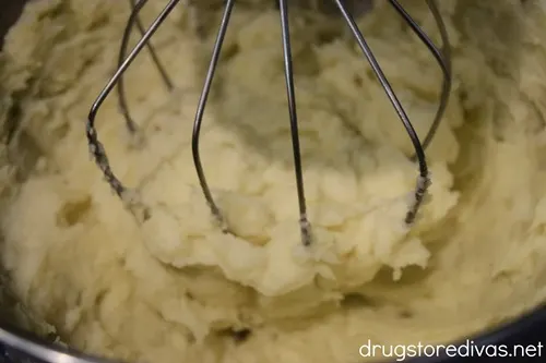 Mashed potatoes in a stand mixer bowl with the whisk attachment stirring them.