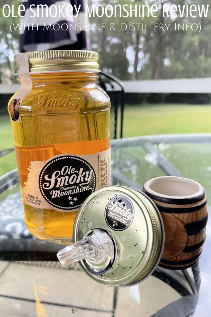 A mason jar of moonshine, a lid with a spout, and a barrel-shaped shot glass on a table with the words "Ole Smoky Moonshine Review with moonshine and distillery info" digitally written on top.