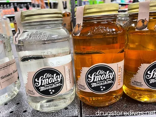 Two bottles of mason jars filled with Ole Smoky Moonshine on a shelf. The one on the left is a clear liquid and the one on the right is a brown liquid.