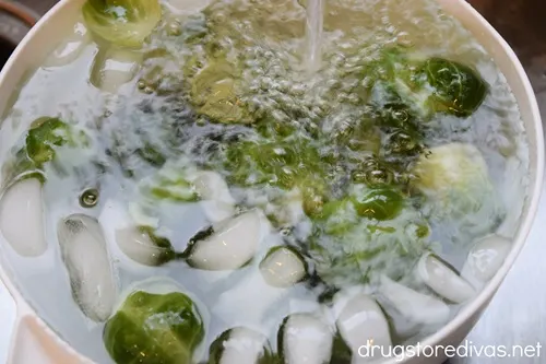 Brussels sprouts in a white bowl with ice cubes and water being poured on them from a faucet. 