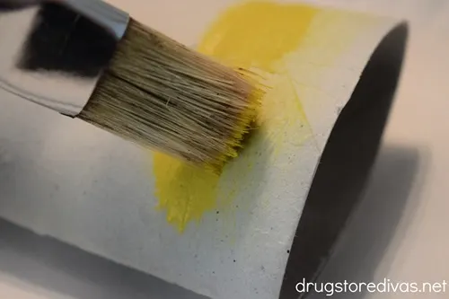 A paint brush painting yellow on a toilet paper roll tube.