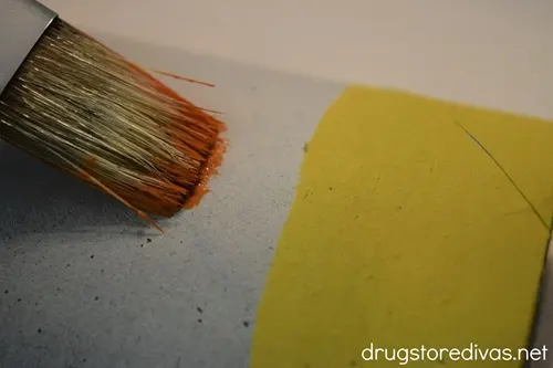 A paint brush painting orange on a toilet paper roll tube that already has a strip of yellow painted.