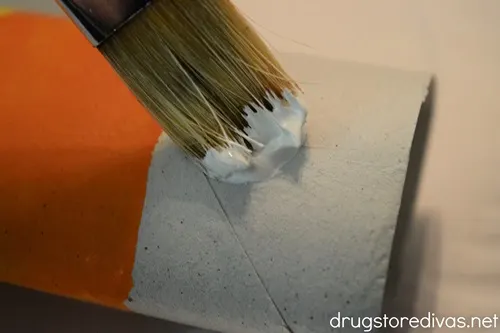A paint brush painting yellow on a toilet paper roll tube that already has a strip of orange painted.