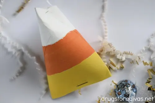 A cardboard tube painted to look like a piece of candy corn on a white tray with white and gold paper shred and a blue Hershey's Kiss near it.