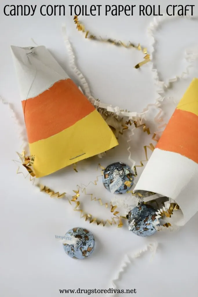 Two toilet paper rolls made to look like candy corn with paper shred and blue Hershey's Kisses on a white tray with the words "Candy Corn Toilet Paper Roll Craft" digitally written on top.