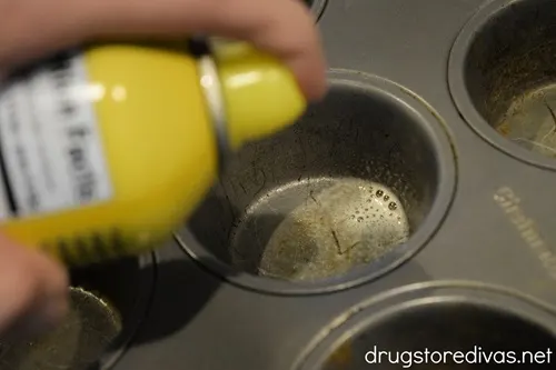 A muffin tin being sprayed with cooking spray.
