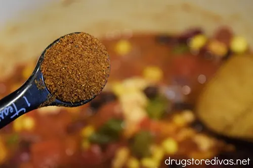 A measuring spoon with a burnt orange seasoning being added to chicken chili.