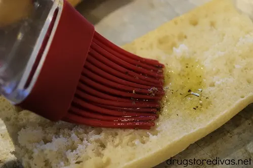 A red pastry brush brushing seasoned olive oil onto French bread.