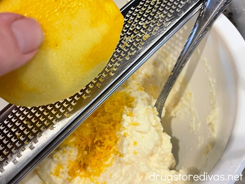 A lemon being zested into a bowl of ricotta cheese that has a silver spoon in it.