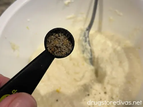 A measuring spoon with spices in it above a bowl with ricotta cheese in it.