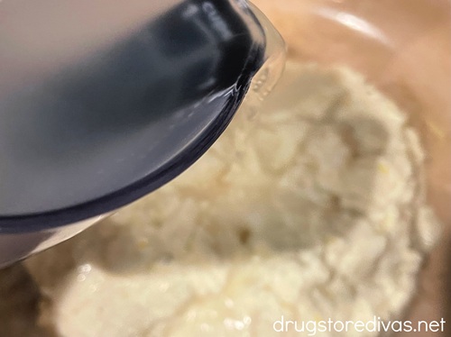 Water in a measuring cup being poured into a bowl with a ricotta cheese mixture in it.