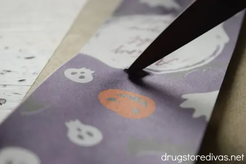 A pair of scissors cutting a slit in a Halloween themed piece of paper.