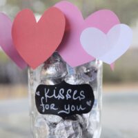 A Hershey's Kisses filled mason jar with paper hearts around the top at and the words 