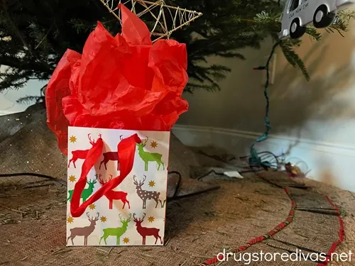 A Christmas gift bag with red and green deer on it and red tissue paper out the top under a Christmas tree.