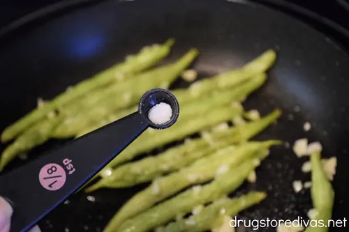 Salt in a measuring spoon above green beans in a skillet.