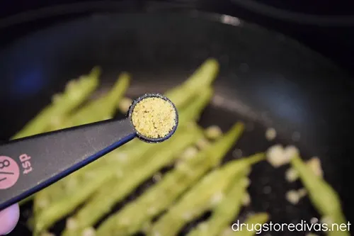 A measuring spoon with spices above green beans in a skillet.