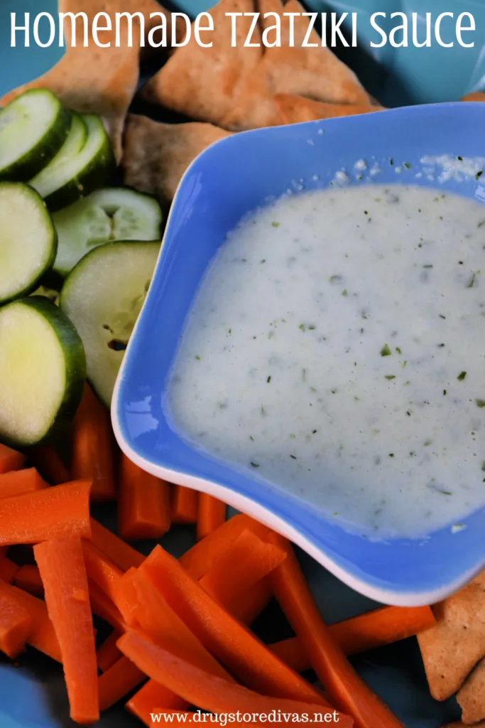 Tzatziki sauce in a blue bowl surrounded by cucumber slices, carrot sticks, and pita chips and the words "Homemade Tzatziki Sauce" digitally written on top.