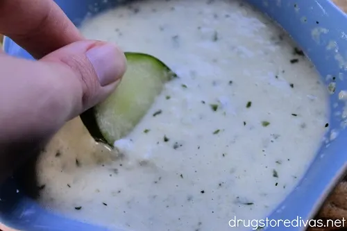 A hand dipping a cucumber slice into tzatziki sauce.