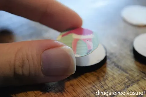 Two fingers holding a round cutout that has a flamingo in a Christmas hat on it over a round, wooden slice.