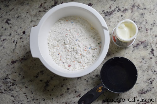 Funfetti cake mix in a bowl next to a container of yogurt and a cup of water.