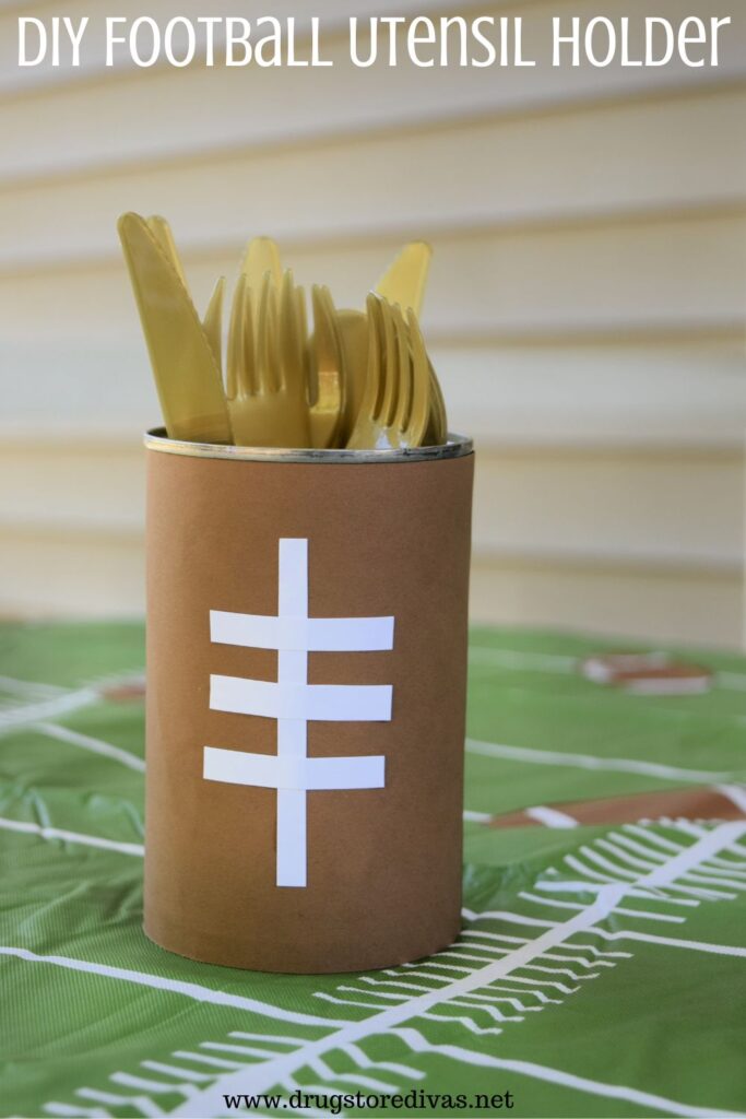 A can, decorated to look like a football, with gold flatware in it, on a tablecloth that looks like a football field with the words "DIY Football Utensil Holder" digitally written on top.