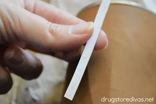 A hand holding a white strip of card stock over a tin can that's wrapped in brown card stock.