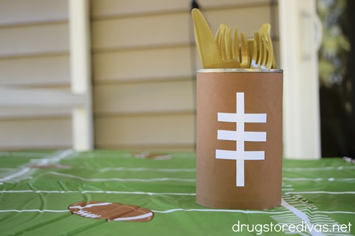 A DIY Football Utensil Holder, which is a can decorated to look like a football, with gold flatware in it, on a tablecloth that looks like a football field.