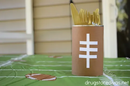 A DIY Football Utensil Holder, which is a can decorated to look like a football, with gold flatware in it, on a tablecloth that looks like a football field.