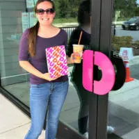 A woman holding a Dunkin' coffee and bag in front of a Dunkin' with the words 