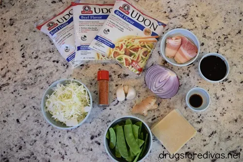 Three packs of Udon noodles, bacon, shredded cabbage, togarashi, garlic, red onion, ginger, snap peas, wonton wrappers, and sauce on a table.