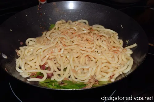 Seasoning being added to cooked udon noodles in a wok on top of vegetables.