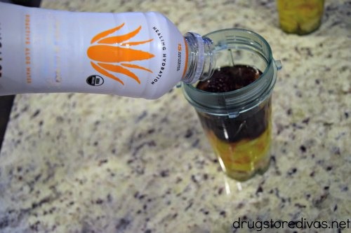 A bottled drink being poured into a blender cup with fruit in it.