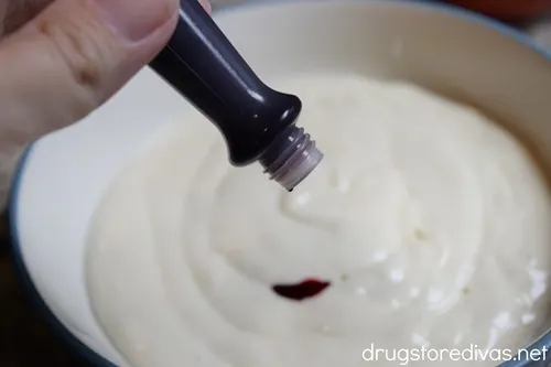A tube of food coloring in a hand being held over a bowl with white cake batter in it.