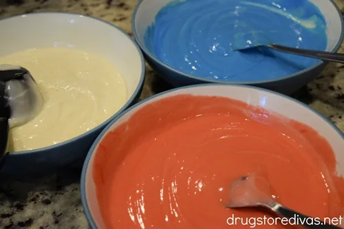 Three bowls, one with white cake batter, one with blue cake batter and a spoon, and one with red cake batter and a spoon.