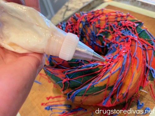 A piping bag filled with white chocolate being held by a hand over a red, white, and blue Bundt cake.