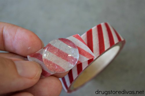 Fingers holding red and white striped tape under a clear flat marble. 