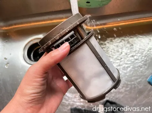 A hand holding a dishwasher filter in a sink with water pouring on it.