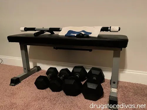 A workout bench with a bar and towel on top and four weights under it.
