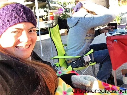 A woman and man sitting at a booth at a farmers market and the woman is wearing a black jacket and purple crocheted headband taking a selfie while the man is in a gray sweatshirt and using a laptop.