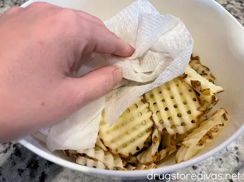 A hand holding a paper towel that's on top of raw waffle fries in a white bowl.