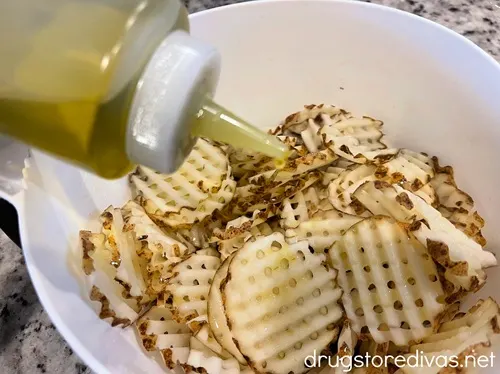 A hand holding a squeeze bottle of oil over raw waffle fries in a white bowl.