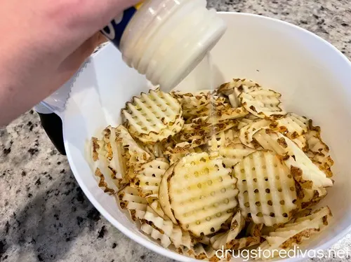 A hand holding a bottle of seasoning over raw waffle fries in a white bowl.