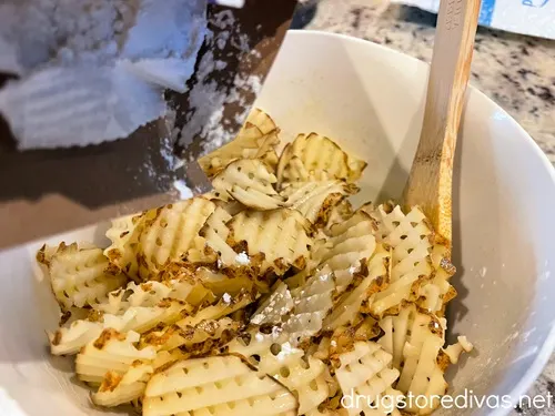 An open box of corn starch being poured over raw waffle fries in a white bowl with a wood spoon in the bowl.