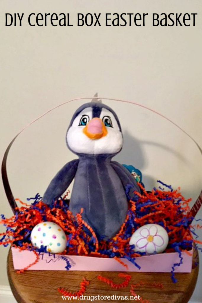 A penguin stuffed animal, paper shred, and eggs in a homemade Easter basket with the words "DIY Cereal Box Easter Basket" digitally written on top.