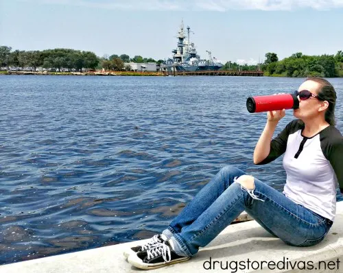 A woman drinking from a travel mug in front of a battleship.