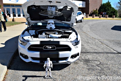 A black and white Mustang car with its hood open, a stormtrooper from Star Wars mask under the hood and a stormtrooper figure in front.