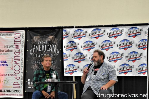Two men at a table, one with a microphone, in front of a banner that says "Up & Coming Weekly", a banner that says, "The All-American Tattoo" and a banner that says "Fayetteville Comic Con."