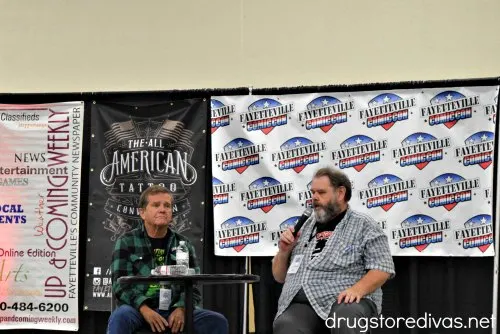 Two men at a table, one with a microphone, in front of a banner that says "Up & Coming Weekly", a banner that says, "The All-American Tattoo" and a banner that says "Fayetteville Comic Con."