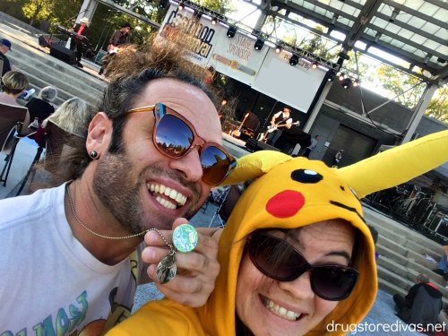 A woman in a Pikachu hoodie next to a man, both are in sunglasses, in front of an outdoor music stage.