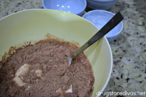 Chocolate cake mix, yogurt, and water in a white bowl with a spoon in it and empty bowls behind it.
